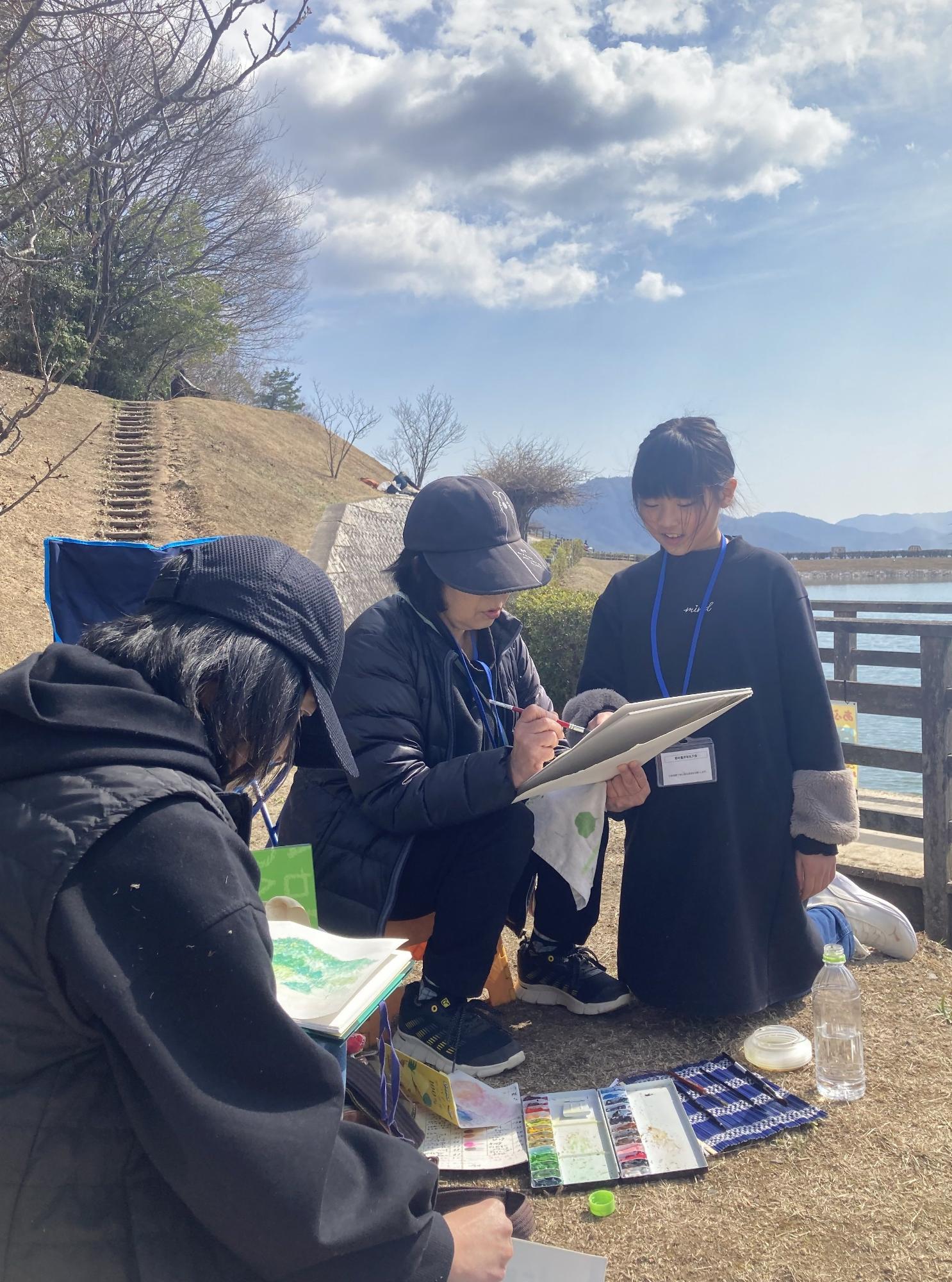 青空のもとで写生大会に参加している女性三名の写真