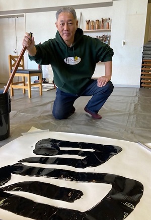 大筆を持ち自分で書いた文字の前でしゃがんでいる緑色のトレーナーの男性の写真