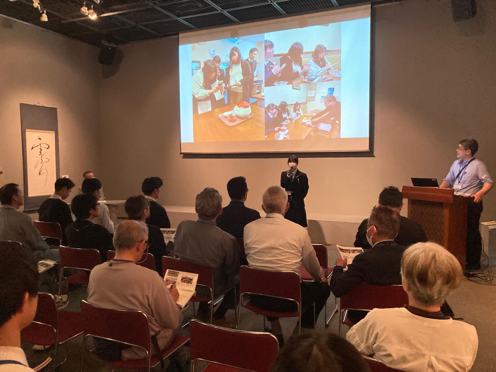 マイパレットラボの報告会でイベント参加の報告をする女子生徒とそれを聞いている参加者の方々の写真