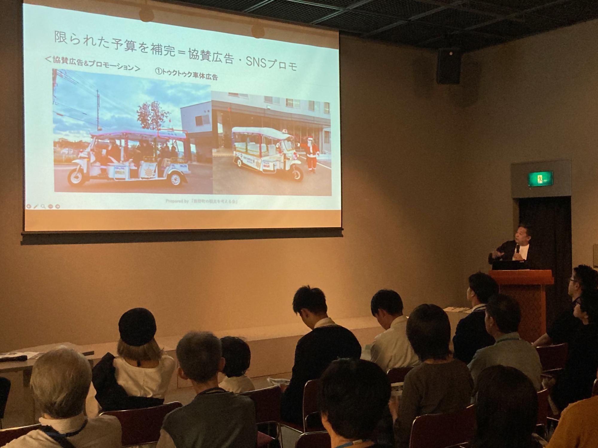 会議でスクリーンに映像を流しながらトゥクトゥク試乗会の活動報告を行う丸山さんとそれを聞いている参加者の方々の写真