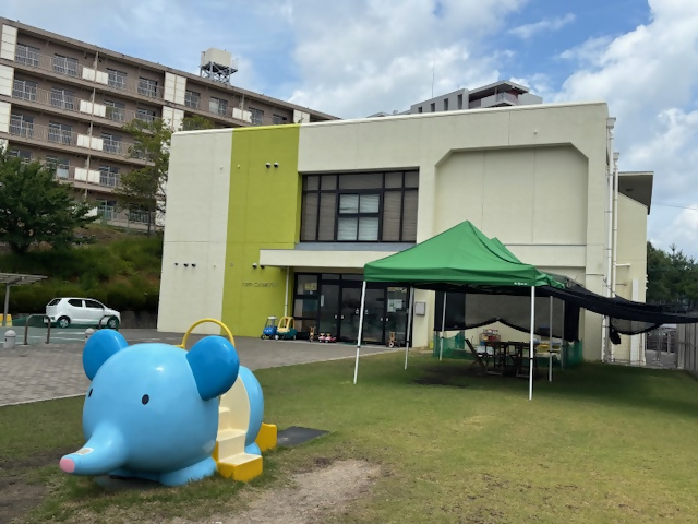 緑色のテントの前に象のすべり台がある子育て支援センターの外観写真